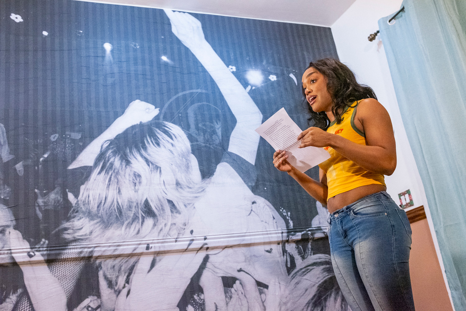 Bailey James reads aloud a short story by Jenn Chand during the third installment of Salad. Behind her to the left, a floor to ceiling, black and white photograph by Gabrielle Ravet shows fans crowdsurfing at a concert.