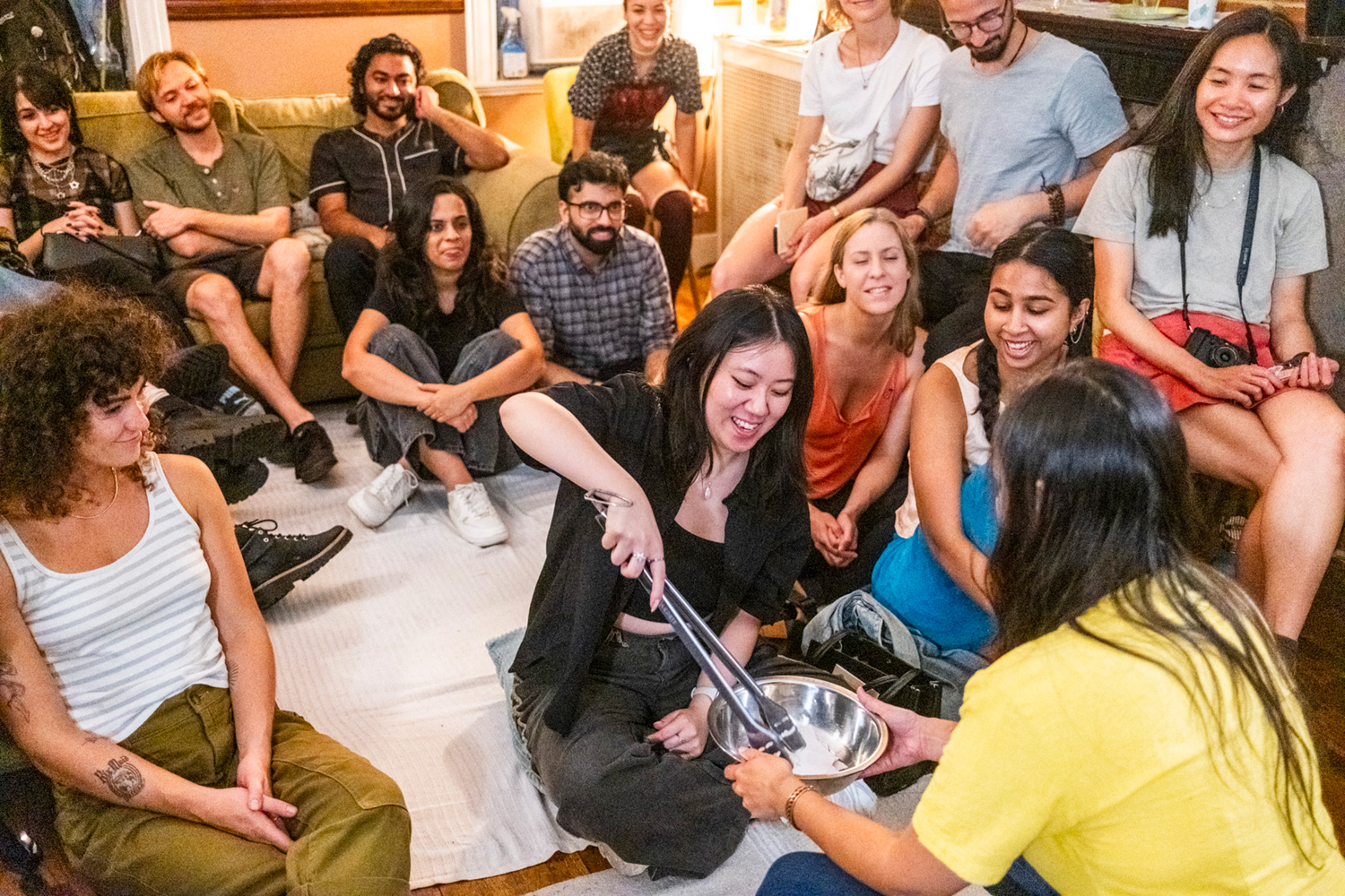 Audience members at the fourth installment of Salad, watch as Chelsea Phou, seated in the front row, picks a slip of paper using oversized metal tongs out of a bowl held by Salad co-creator Sio Tepper.